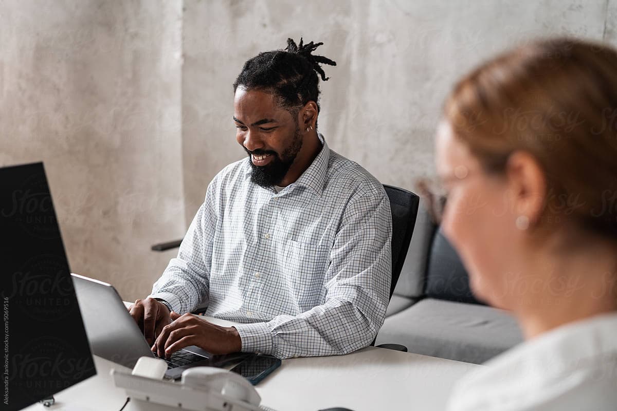 Smiling black man sitting at the desk and working on the laptop computer. His female co-worker is anonymous and blurred.